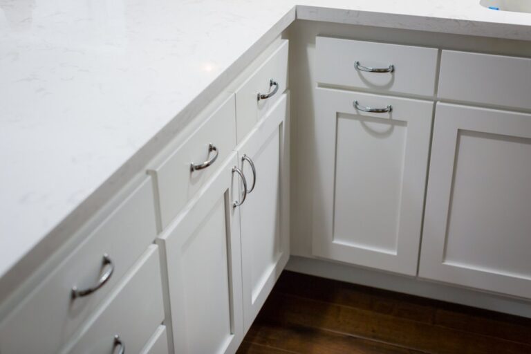 White kitchen cabinets with silver handles and a white marble countertop on a dark wood floor.