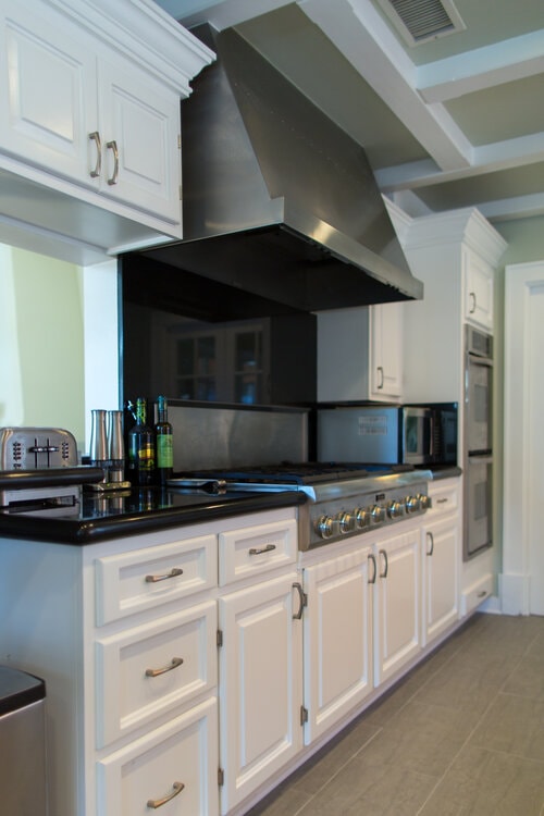 Side view of a remodeled kitchen showcasing freshly refinished white cabinets with brass pulls, black stone countertops, a commercial stainless-steel range hood, and a six-burner gas stove.