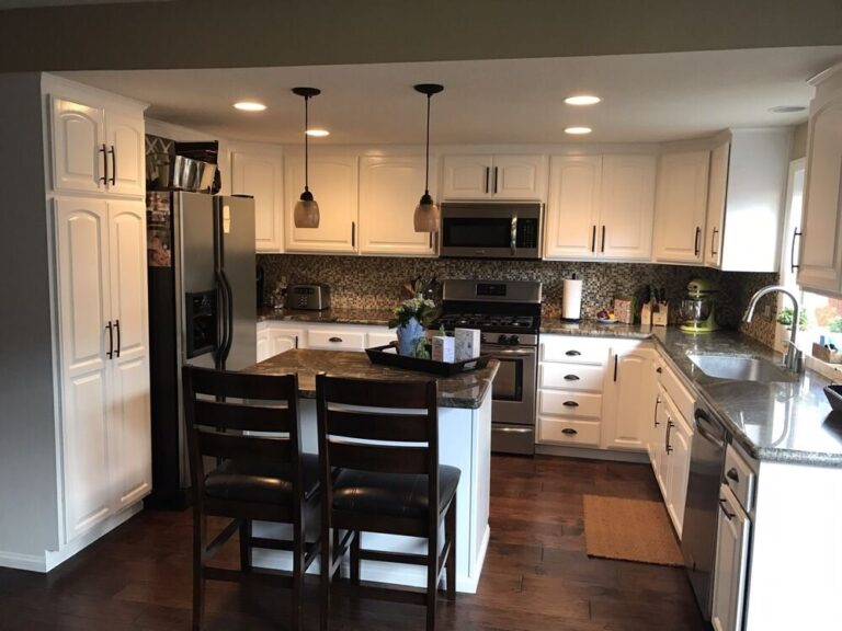 Modern kitchen with white cabinets, stainless steel appliances, dark wood floor, and small island with two chairs.