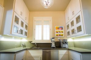 Bright kitchen with white cabinets, a small sink, coffee machines, and colorful art on the wall near a window.