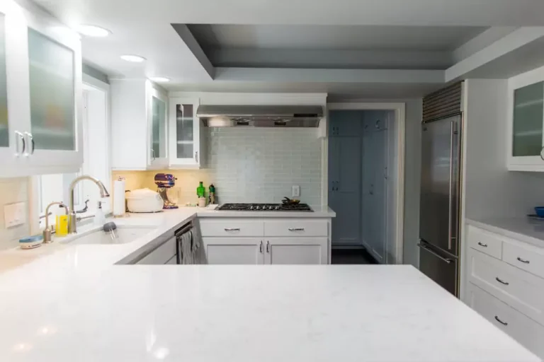 Modern white kitchen with stainless steel appliances, gas stove, and marble countertops, lit by recessed lighting.
