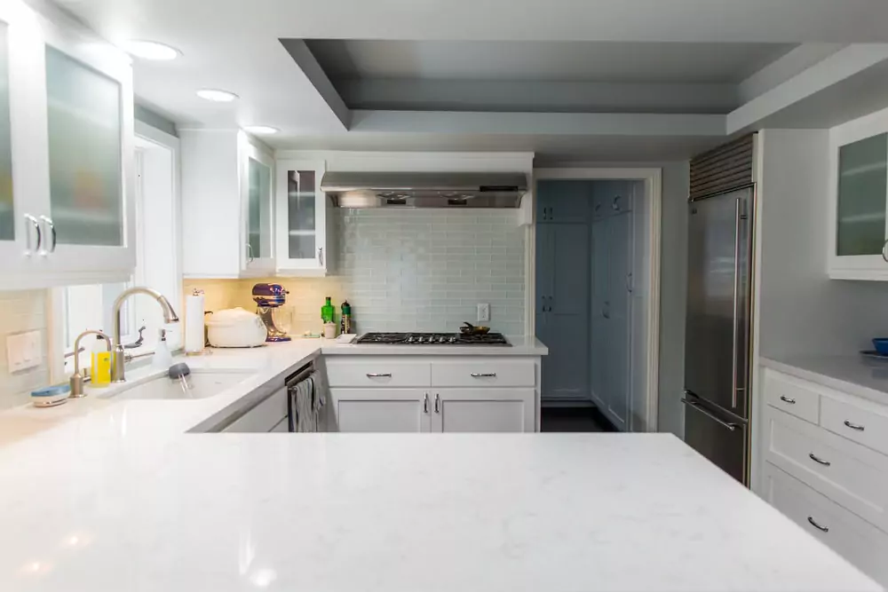 Modern white kitchen with stainless steel appliances, gas stove, and marble countertops, lit by recessed lighting.