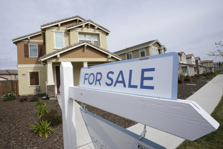 A two-story suburban house with a For Sale sign in the front yard on a clear day.