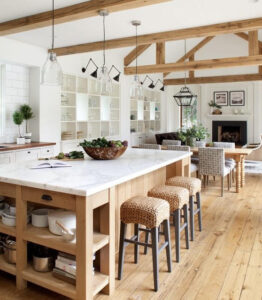 Bright, airy kitchen with wooden beams, marble island, woven stools, open shelves, and dining area with fireplace.