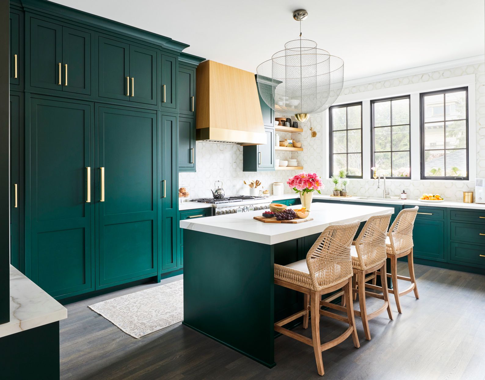 Modern kitchen with dark green cabinets, a white island, wicker barstools, and large windows letting in natural light.