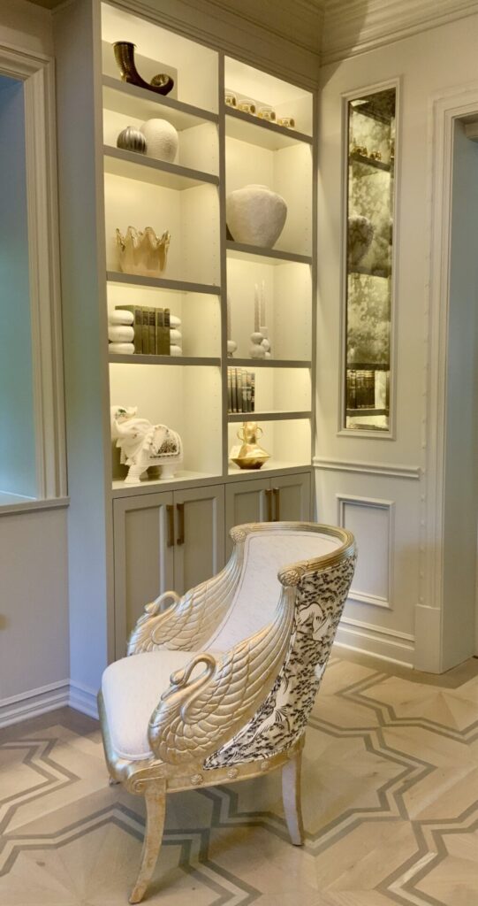 Elegant chair with gold accents sits in front of a built-in shelf displaying decorative objects and books in a stylish room.