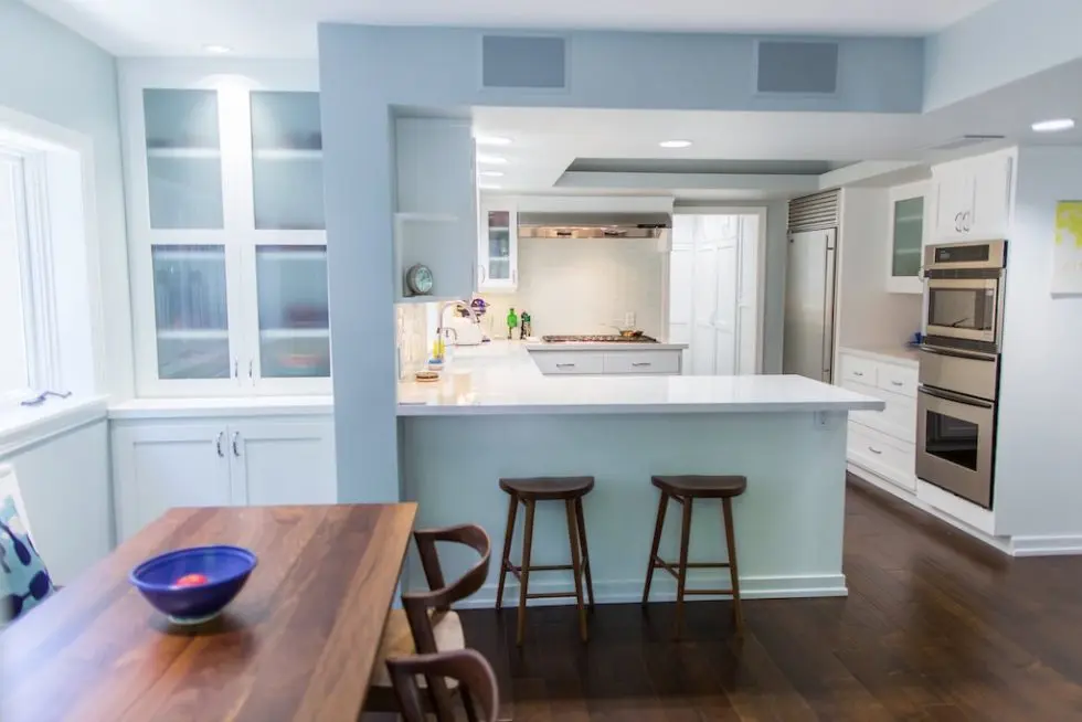 Modern kitchen with white cabinets, stainless steel appliances, island with two stools, and a wooden dining table.