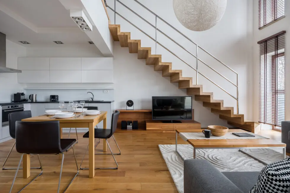 Modern loft apartment with wooden stairs, open kitchen, dining table, TV, and large windows with blinds.