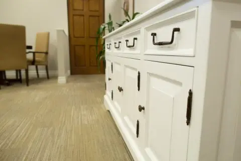 White cabinet with black handles in a carpeted room, with chairs and a wooden door in the background.