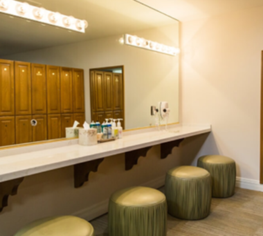 Vanity area with a large mirror, light bulbs, stools, and toiletries; wooden lockers in the background.