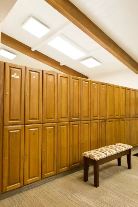 Wooden lockers line the wall of a locker room, with a patterned bench in front on a carpeted floor.