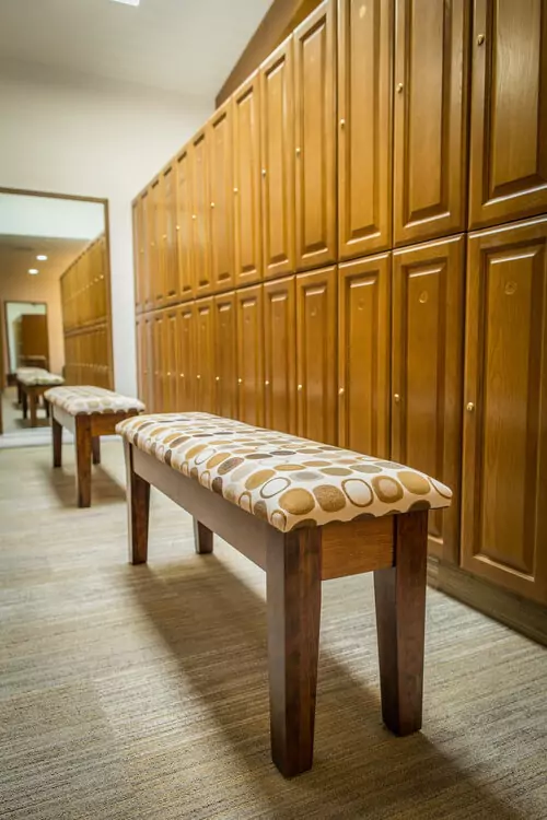 A locker room with wooden lockers and cushioned benches with patterned upholstery on a carpeted floor.