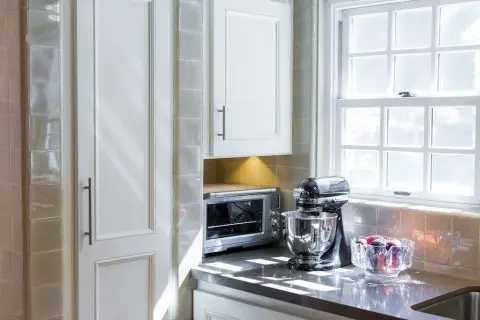 Sunlit kitchen counter with a stand mixer, toaster oven, and fruit bowl beside a freshly stained cabinet near the window.