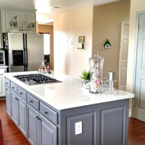 Bright kitchen with a marble island, gray cabinets, stove top, and stainless steel fridge in the background.