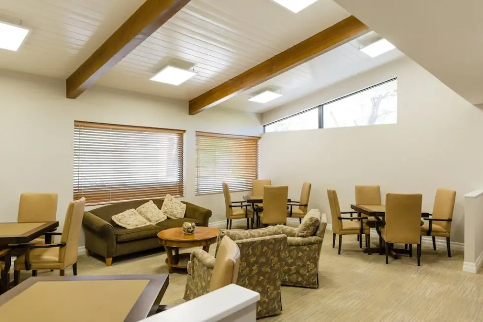 A bright lounge with beige chairs, patterned armchairs, sofas, round tables, and large windows with wooden blinds.