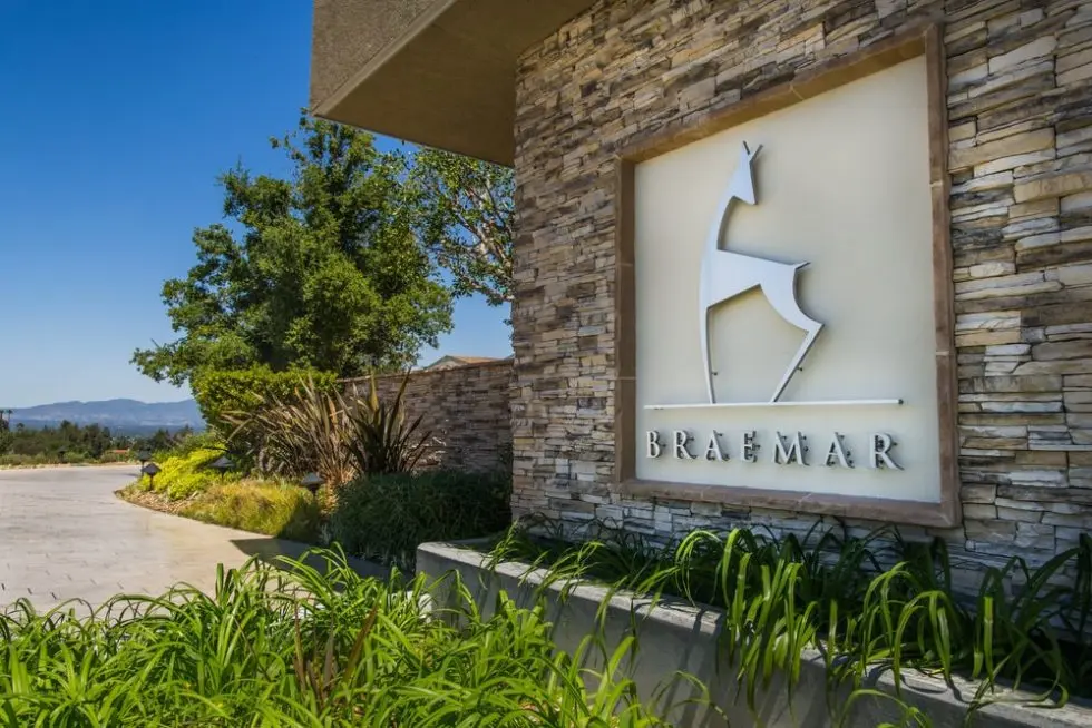 A stone sign for “Braemar” with a stylized deer logo, surrounded by greenery and trees under a clear blue sky.
