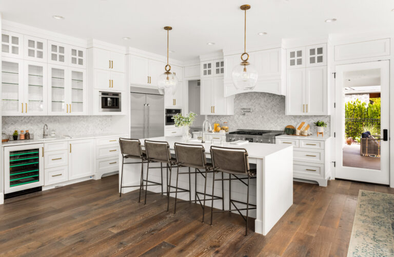Modern white kitchen with island, bar stools, pendant lights, stainless steel appliances, and wooden floors.
