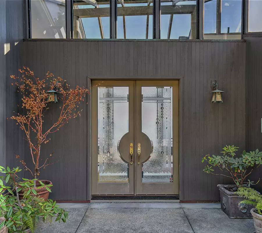 Double glass doors with circular handles, flanked by potted plants and wall lanterns on a modern dark-paneled exterior.