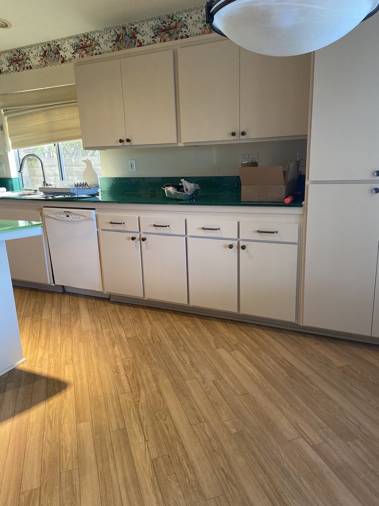 A kitchen with light wood cabinets, green backsplash, wood floor, and sunlight streaming through a window above the sink.