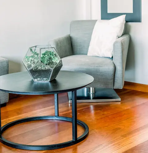 A gray armchair with a white pillow and a glass terrarium on a round black coffee table on a wooden floor.