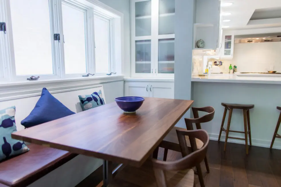 Modern kitchen with wooden table and chairs, window bench with pillows, and white cabinets in the background.