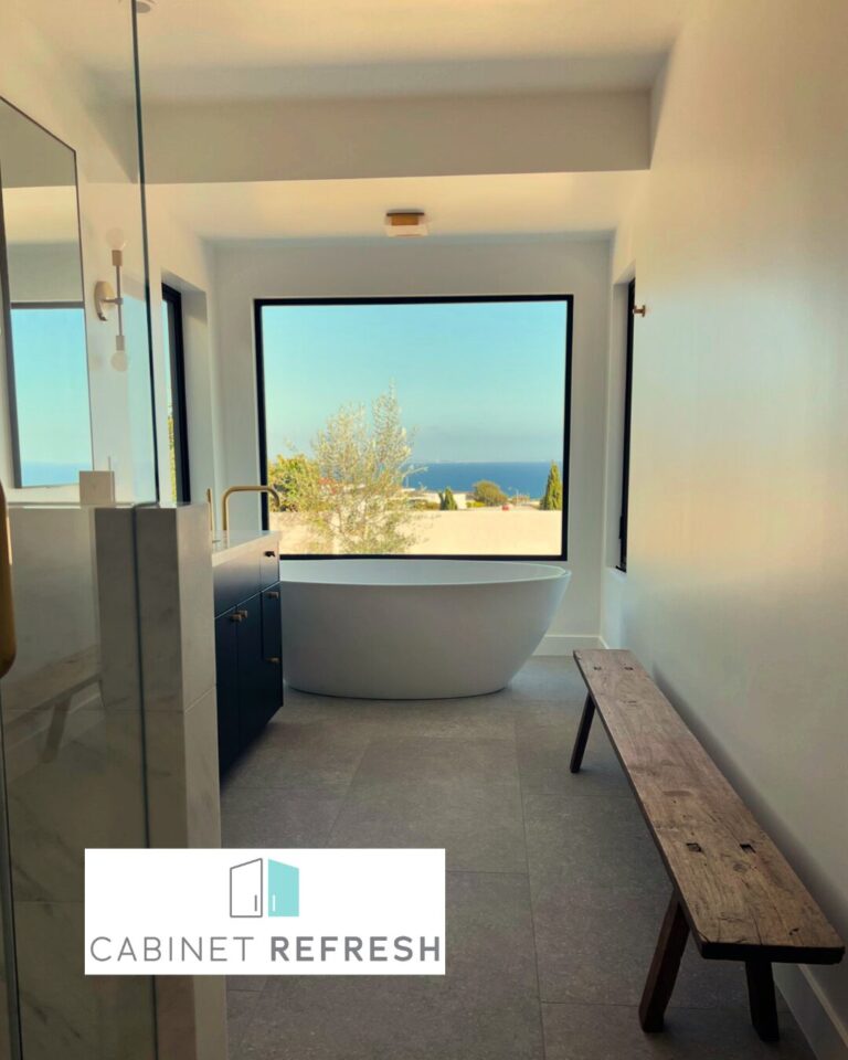 Modern bathroom featuring freestanding bathtub, custom vanity, large picture window, and scenic outdoor ocean view.