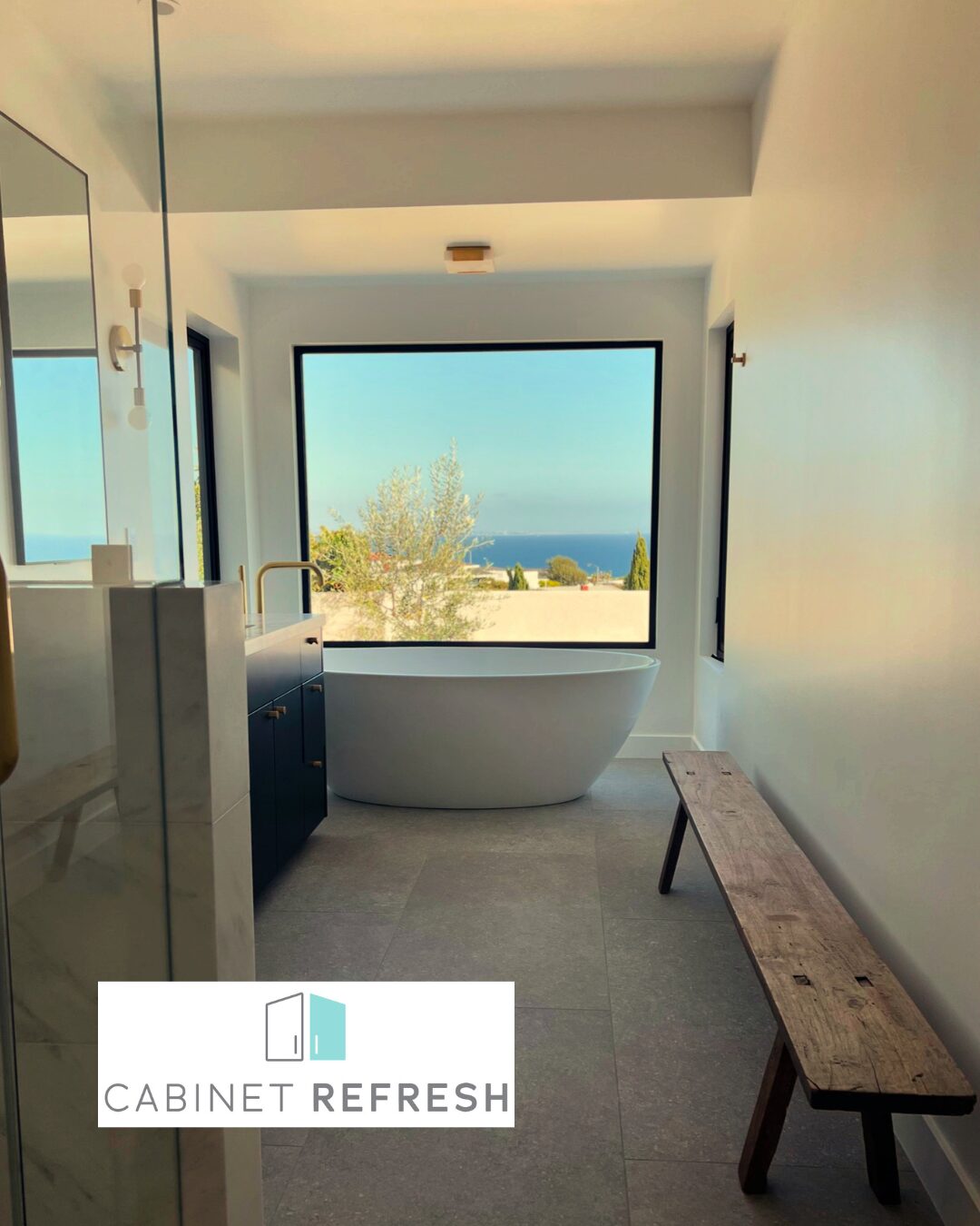 Modern bathroom featuring freestanding bathtub, custom vanity, large picture window, and scenic outdoor ocean view.