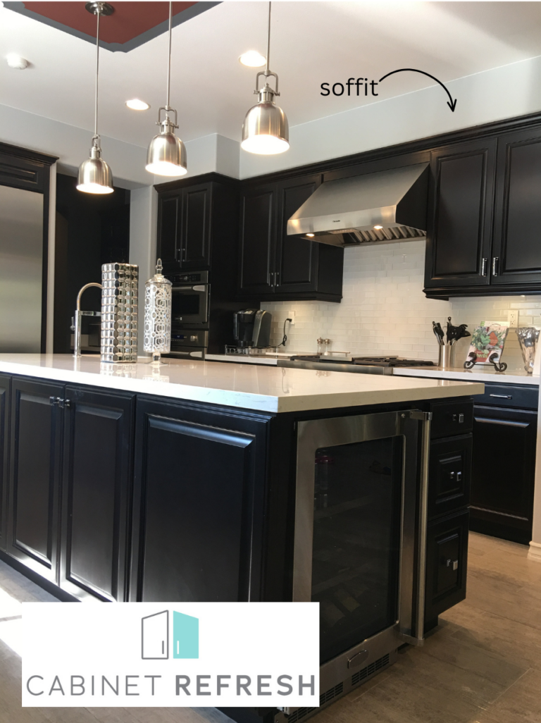 A modern kitchen featuring dark cabinets, a white island countertop, stainless steel appliances, and a boxed-in soffit above the cabinets, highlighted with an arrow.